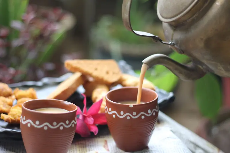 A warm cup of tea being poured from a kettle, surrounded by snacks and a flower