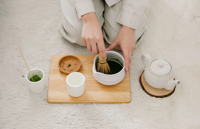 Hands preparing matcha tea with a whisk on a wooden board. Includes teapot and cups