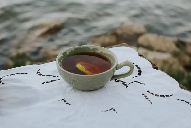 A ceramic cup of tea with a lemon slice, elegantly placed on a lace cloth by a serene lakeside