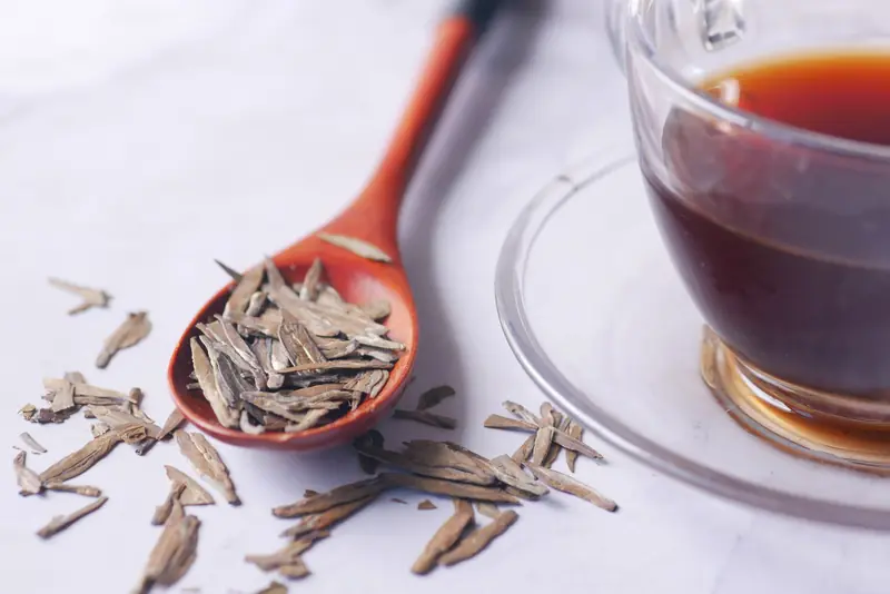 A close-up of dry herbal tea leaves on a wooden spoon next to a cup of brewed tea