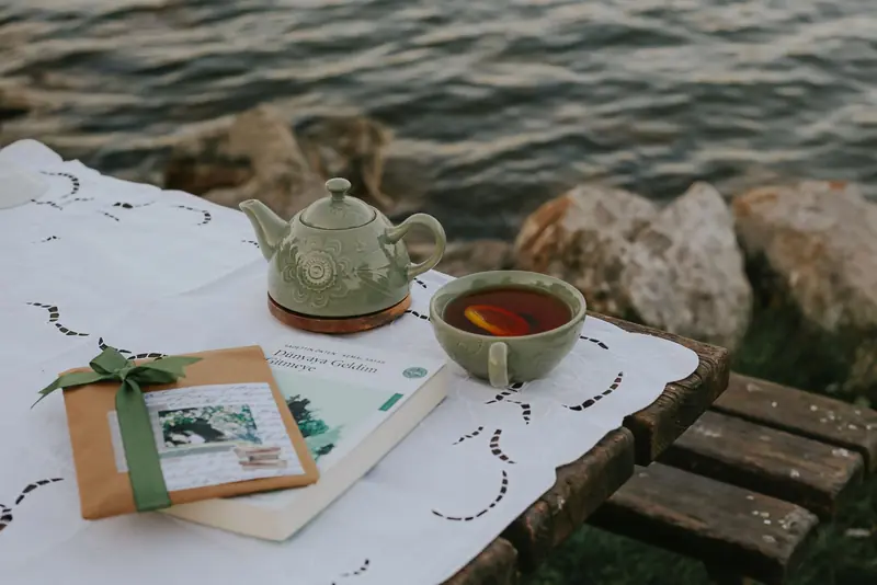 A serene lakeside table with a teapot, cup of tea, book, and wrapped gift on a lace tablecloth