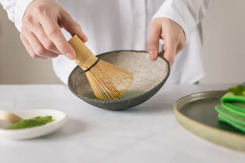 Crop unrecognizable cook in white shirt mixing dry food coloring with water in bowl using whisk on table with green pancakes