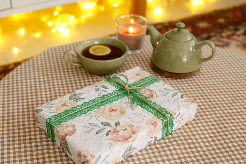 A cozy tea setting with a teapot, cup of tea, candle, and floral-wrapped gift on a checkered tablecloth