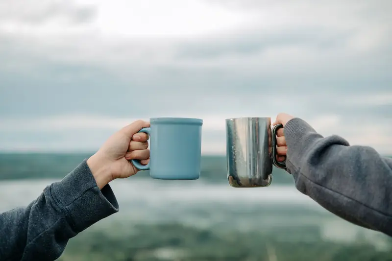 Two hands holding mugs outdoors with a scenic blurry background, symbolizing friendship and relaxation